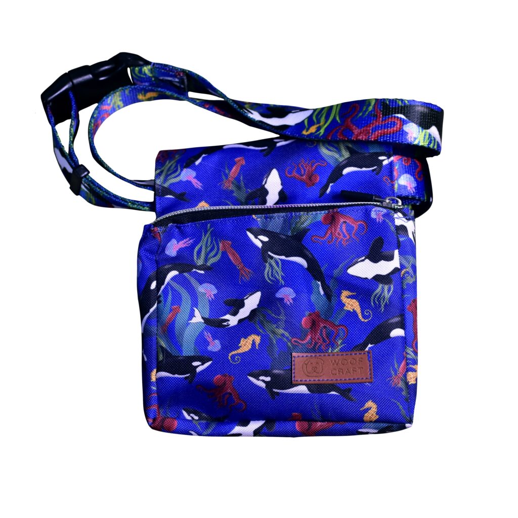Ilustracja przedstawia saszetkę na przysmaki dla psów. Ma ona kolor niebieski i nadruk przedstawiający orki i inne morskie stworzenia.