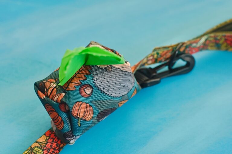 Ilustracja przedstawia ciemnoniebieskie etui na kupoworki dla psa, które jest przymocowane do smyczy. Nadruk na taśmie oraz etui przedstawia jesienne motywy: jeże, żołędzie i liście.