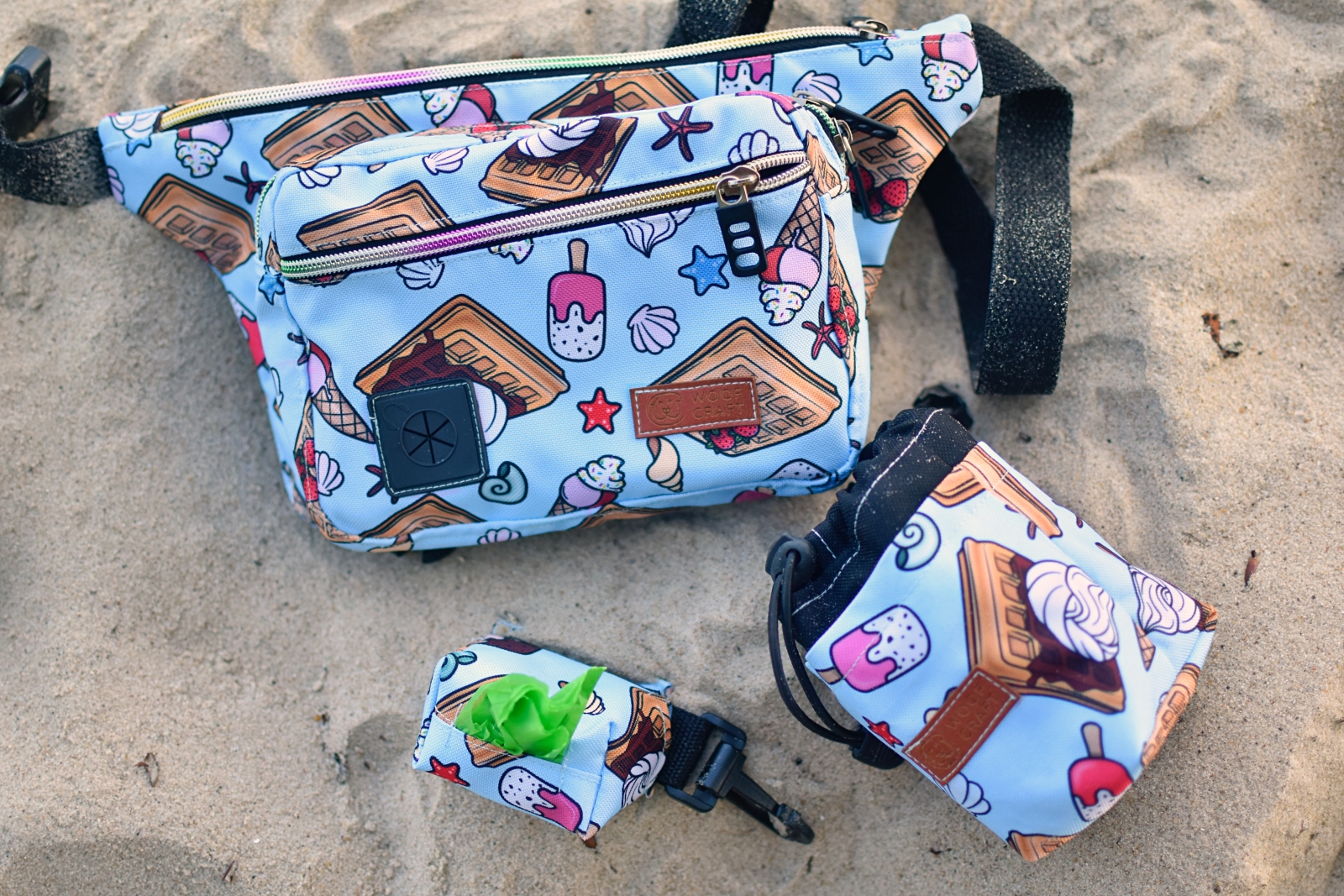 Ilustracja przedstawia akcesoria spacerowe w kolorze niebieskim. Nerka spacerowa. Saszetka na przysmaki dla psa. Etui na kupoworki. Akcesoria leżą na plaży.